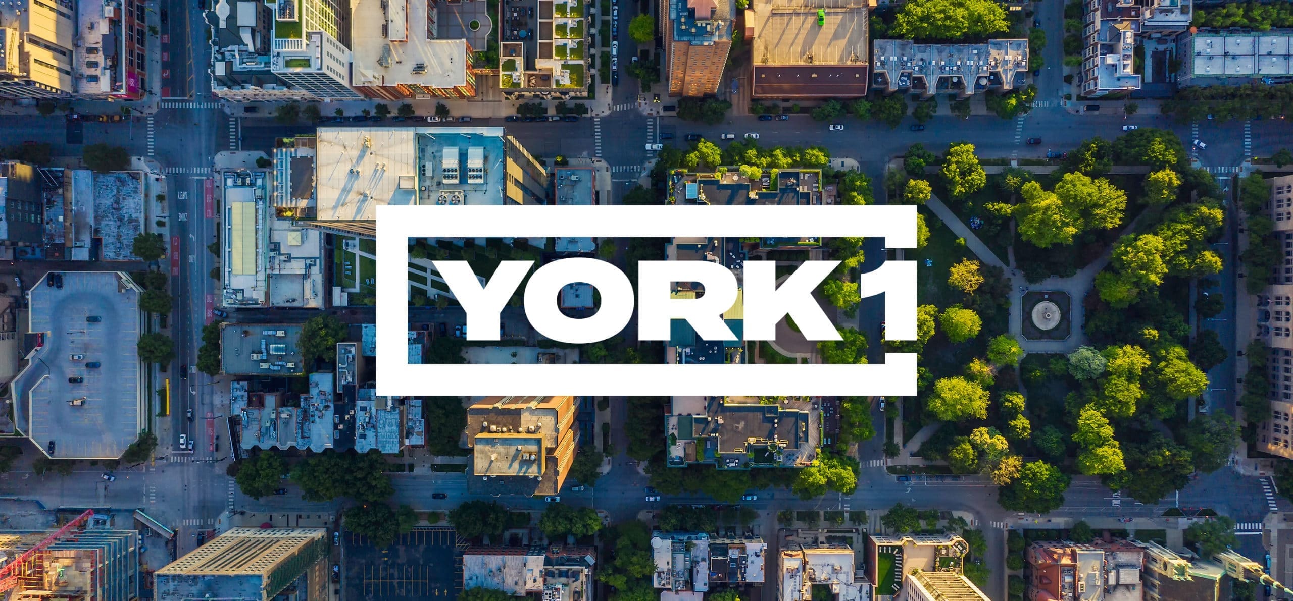 Bird's-eye view of cityscape. Buildings, busy streets, trees, and a park. White York1 logo in the centre.