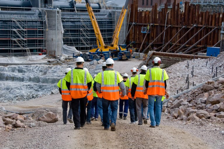 Rear shot of a group of construction workers on a job site. They are walking down a gravel hill towards machinery and construction project