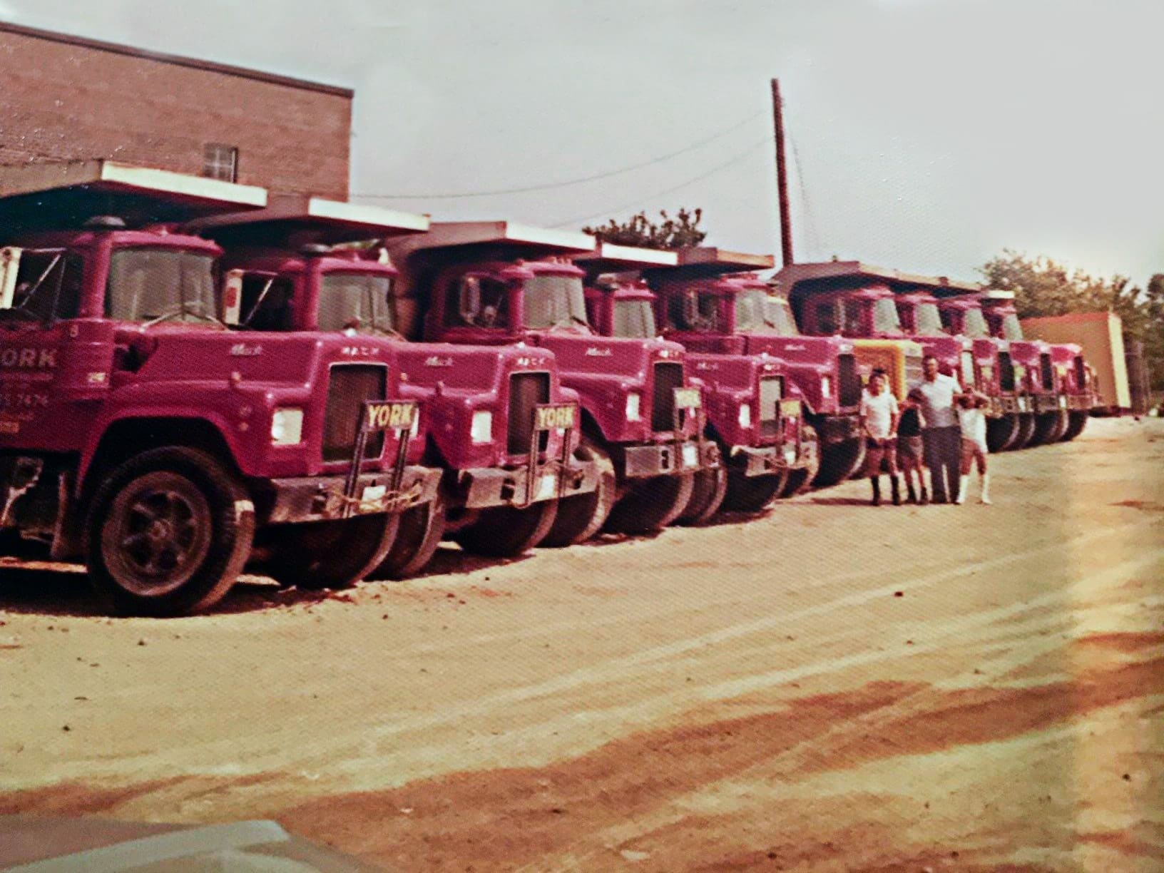 Vintage style photo. Row of red York trucks parked on a dirt driveway with Santo, York's founder, and his three sons standing in front.
