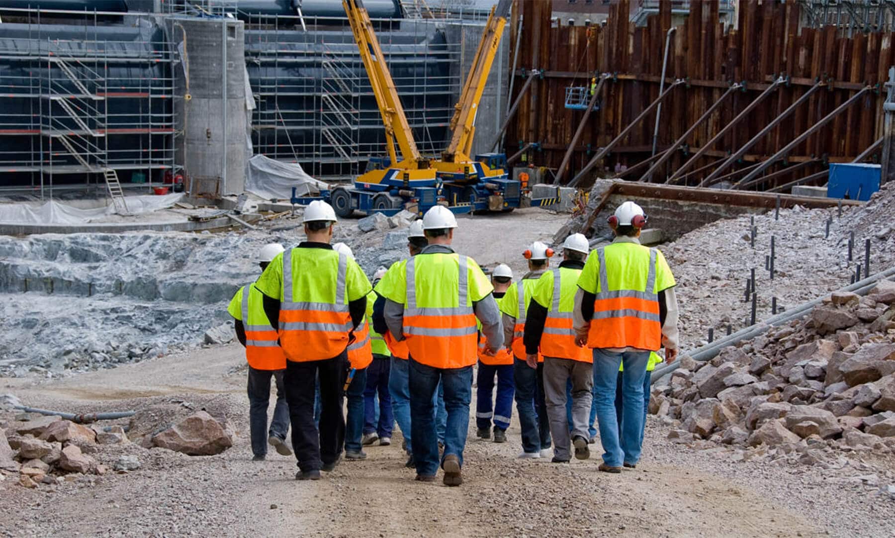Rear shot of a group of construction workers on a job site. They are walking down a gravel hill towards machinery and a construction project.