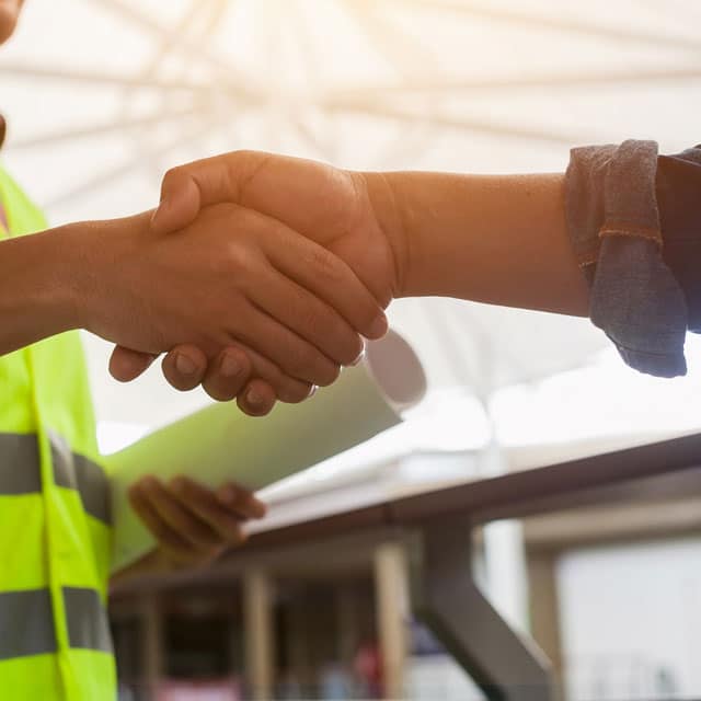 Shaking hands. One person holding rolled-up construction plans wearing yellow hi-vis vest, other person's arm in denim shirt.