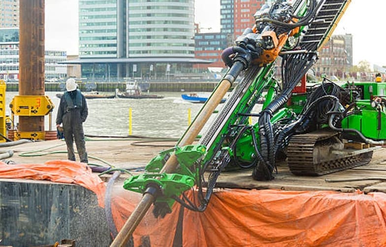Green directional drill on site. Worker beside drill and city buildings in background