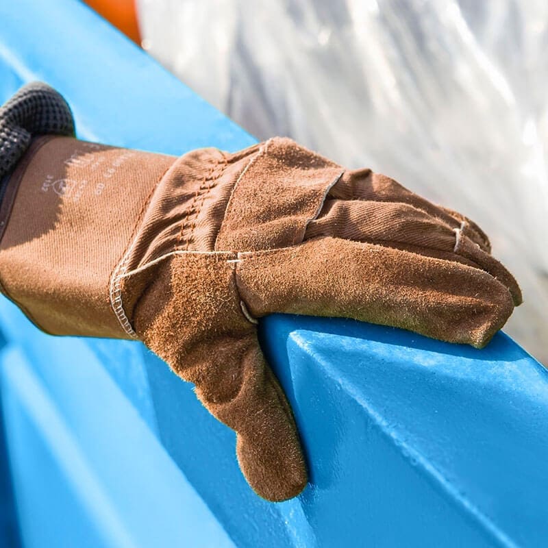 A hand wearing brown work glove grabbing the top of a blue roll-off bin