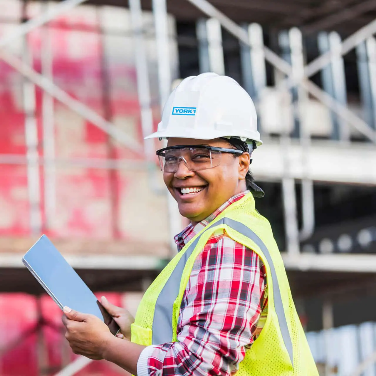 Smiling worker on job site carrying a tablet. Scaffolding in the background. Wearing white York1 hardhat, clear goggles, and yellow hi-vis vest.