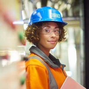 Smiling worker carrying a tablet. Wearing blue York1 hardhat, clear goggles and orange hi-vis jacket.