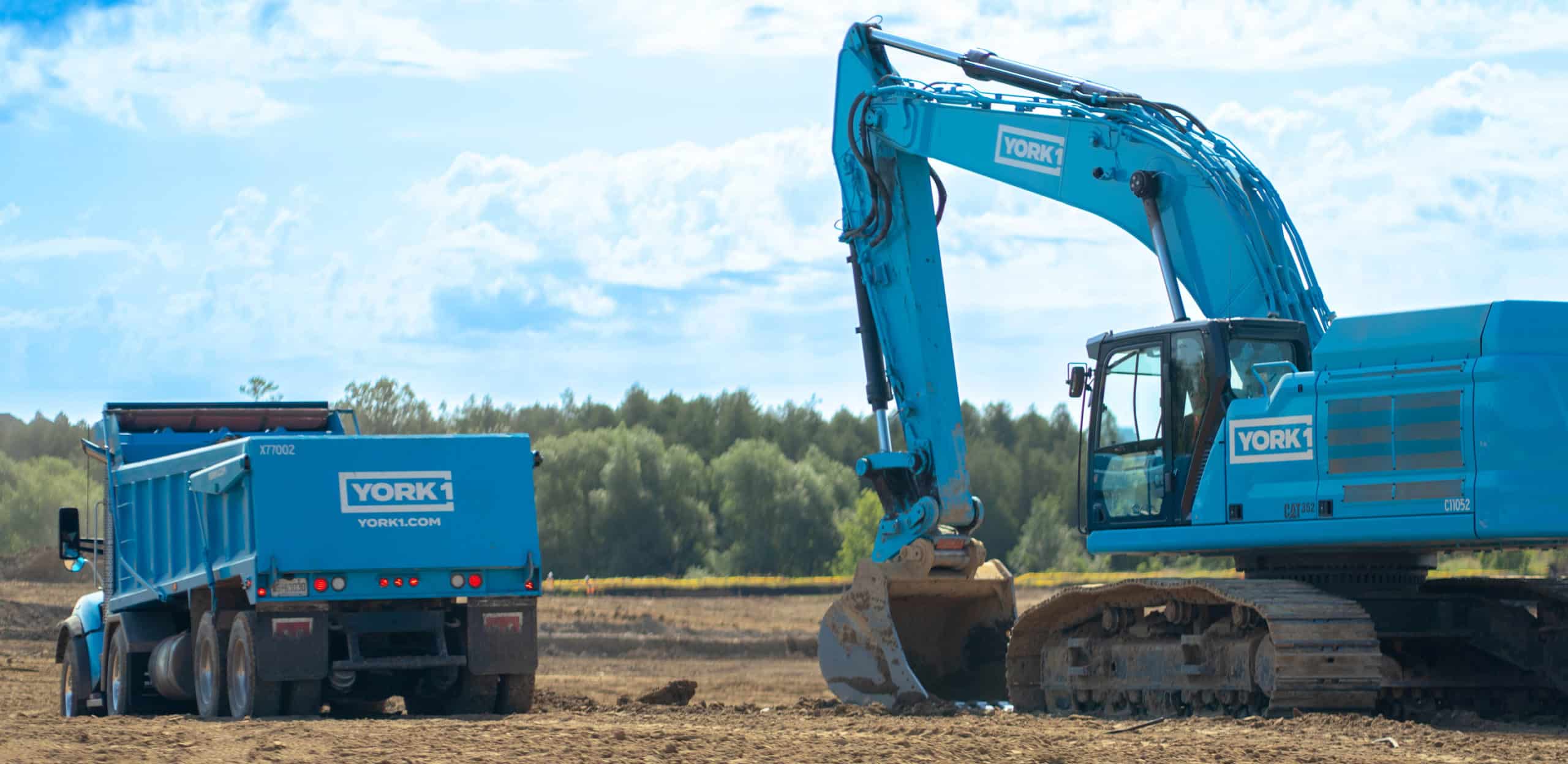 A cyan York1 excavator is loading dirt into a York1 dump truck in an open field with trees in the background.