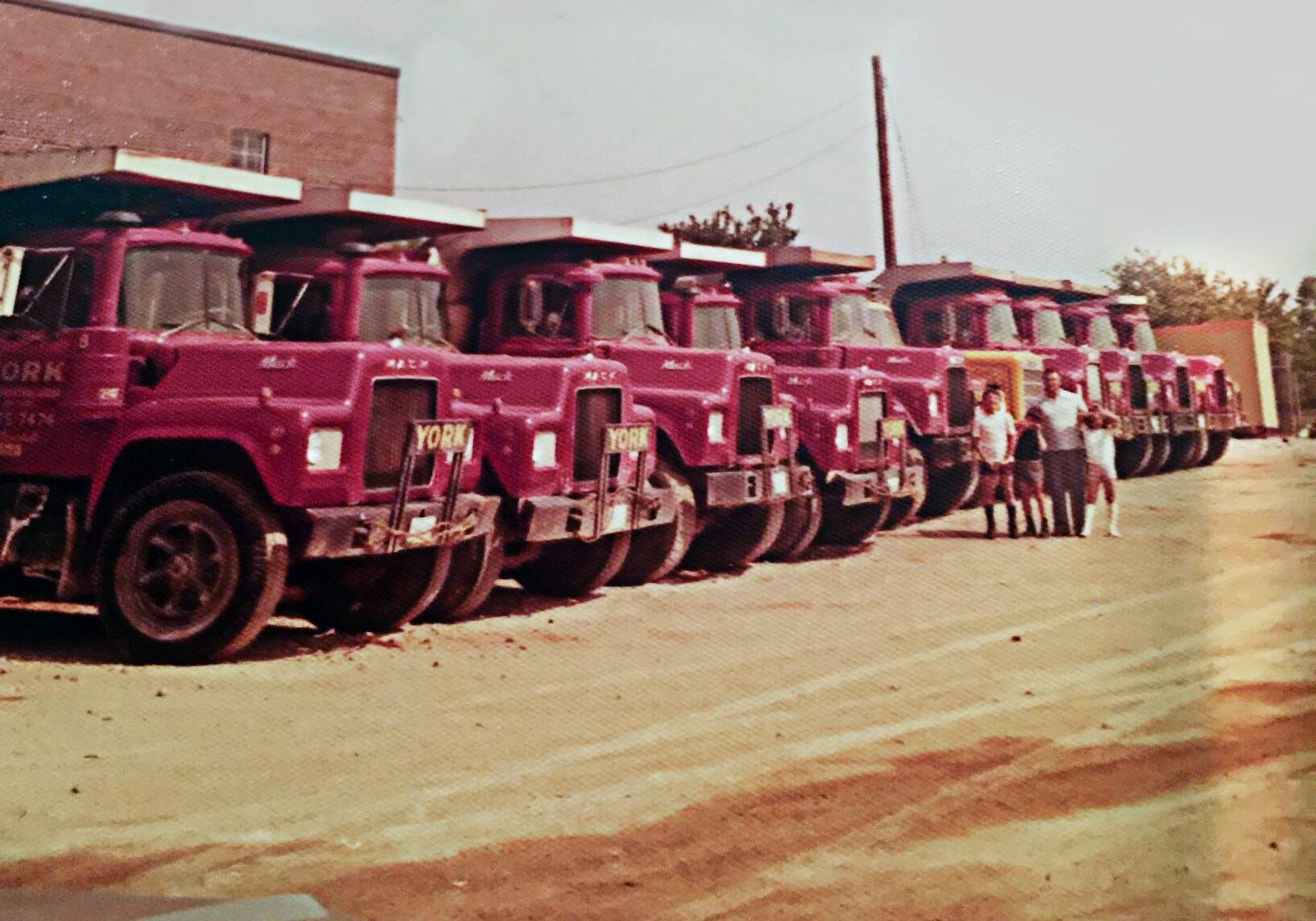 Vintage style photo. Row of red York trucks parked on a dirt driveway with Santo, York's founder, and his three sons standing in front.