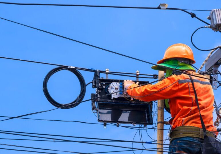 Rear view of technician on wooden ladder is working to install fiber optic and splitter box on electric pole against blue sky background